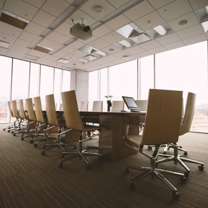 A number of rolling chairs line a long table in an empty meeting room with floor to ceiling windows.